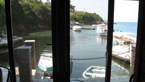 Blick aus einem Zimmer mit Glastür auf einen Yachthafen, wo Boote vor Anker liegen, eine Steinmauer und ein kleiner Tisch sind zu sehen.