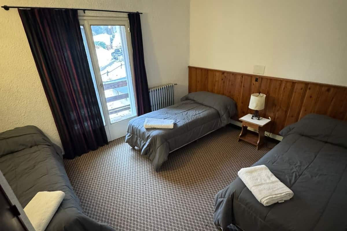 Habitación de hotel con dos camas individuales, una mesita con lámpara y un ventanal con cortinas.