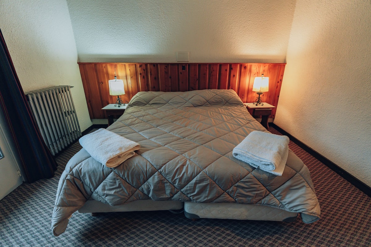 Una cama bien hecha con una colcha acolchada y dos toallas, cabecera de madera, dos lámparas de noche y un radiador.