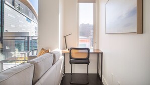 A modern living room with a sofa, a desk, and a chair. A large window offers a view of a building with the sign "INSERVO". - Modern Studio Apt with Rooftop, Gym, and Parking (Milwaukee)