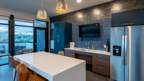 A modern kitchen with a large island, dark blue cabinets, stainless steel appliances, and a city view through large windows. - Modern Studio Apt with Rooftop, Gym, and Parking (Milwaukee)