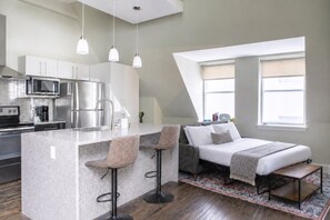 A modern kitchen with stainless steel appliances, a central island with bar stools, a bed with a view, and a large window. - Downtown Luxury 1BR Loft · Walkable · Sleeps 4 (Milwaukee)