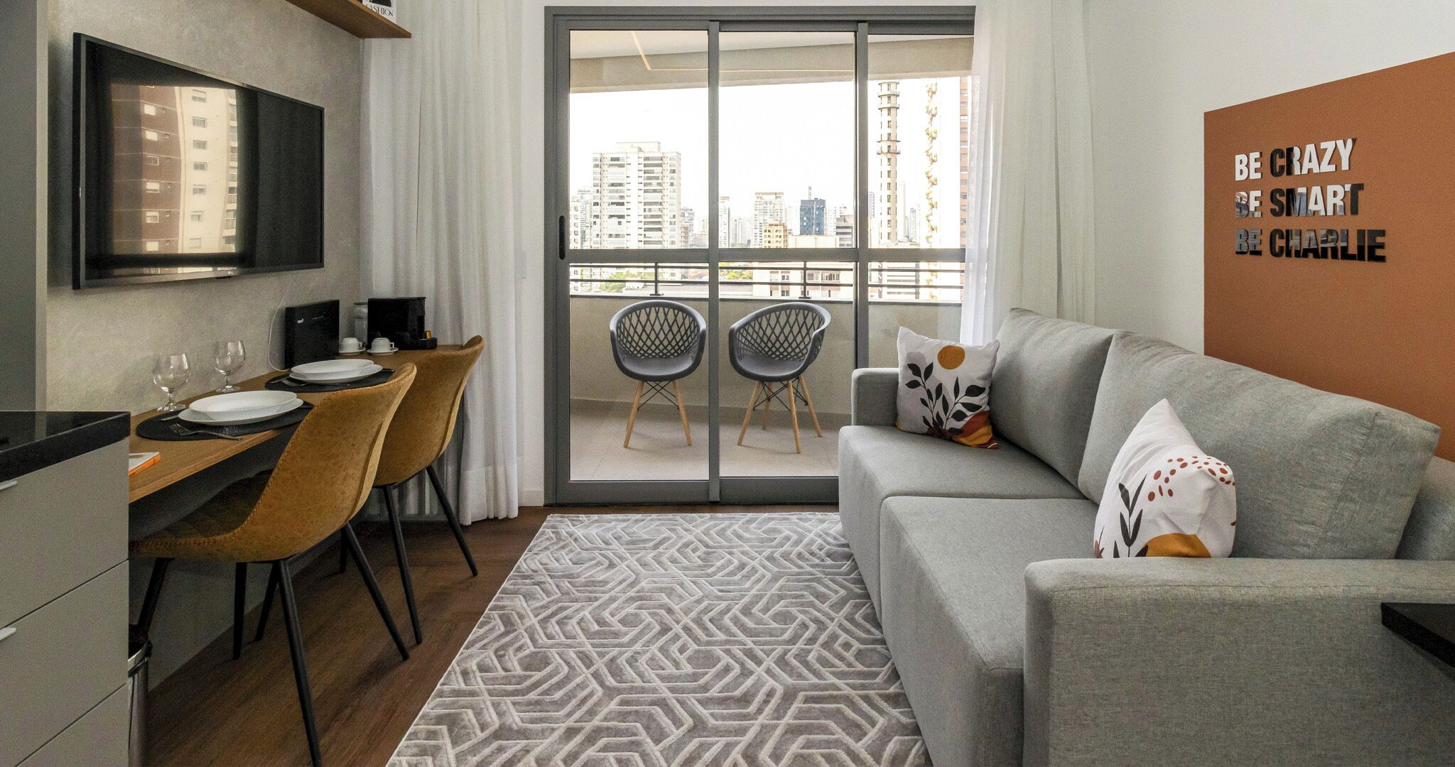 A modern living room with a grey sofa, a dining area with two chairs, and a view of high-rise buildings through a glass door.