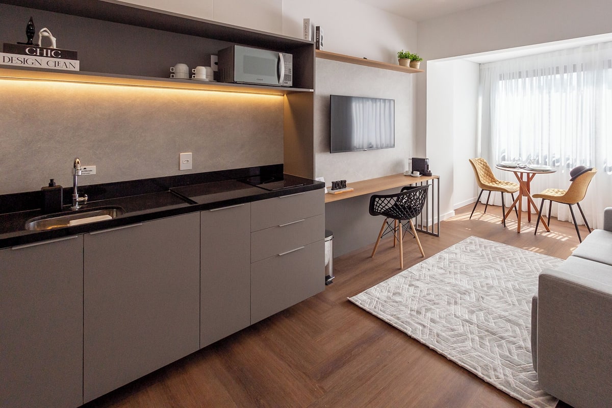 Eine moderne Küche mit Mikrowelle, Kaffeemaschine und einem Fernseher über der Arbeitsfläche. Es gibt einen Essbereich mit Tisch und Stühlen sowie eine Couch mit einem gemusterten Teppich.