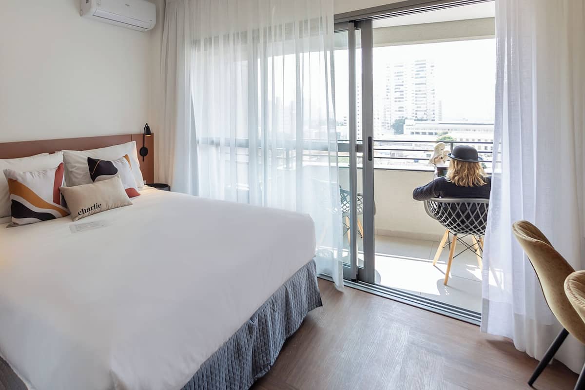 Ein Hotelzimmer mit einem großen Bett, Blick auf die Stadt durch eine Schiebetür und einem Balkon mit einem Stuhl.