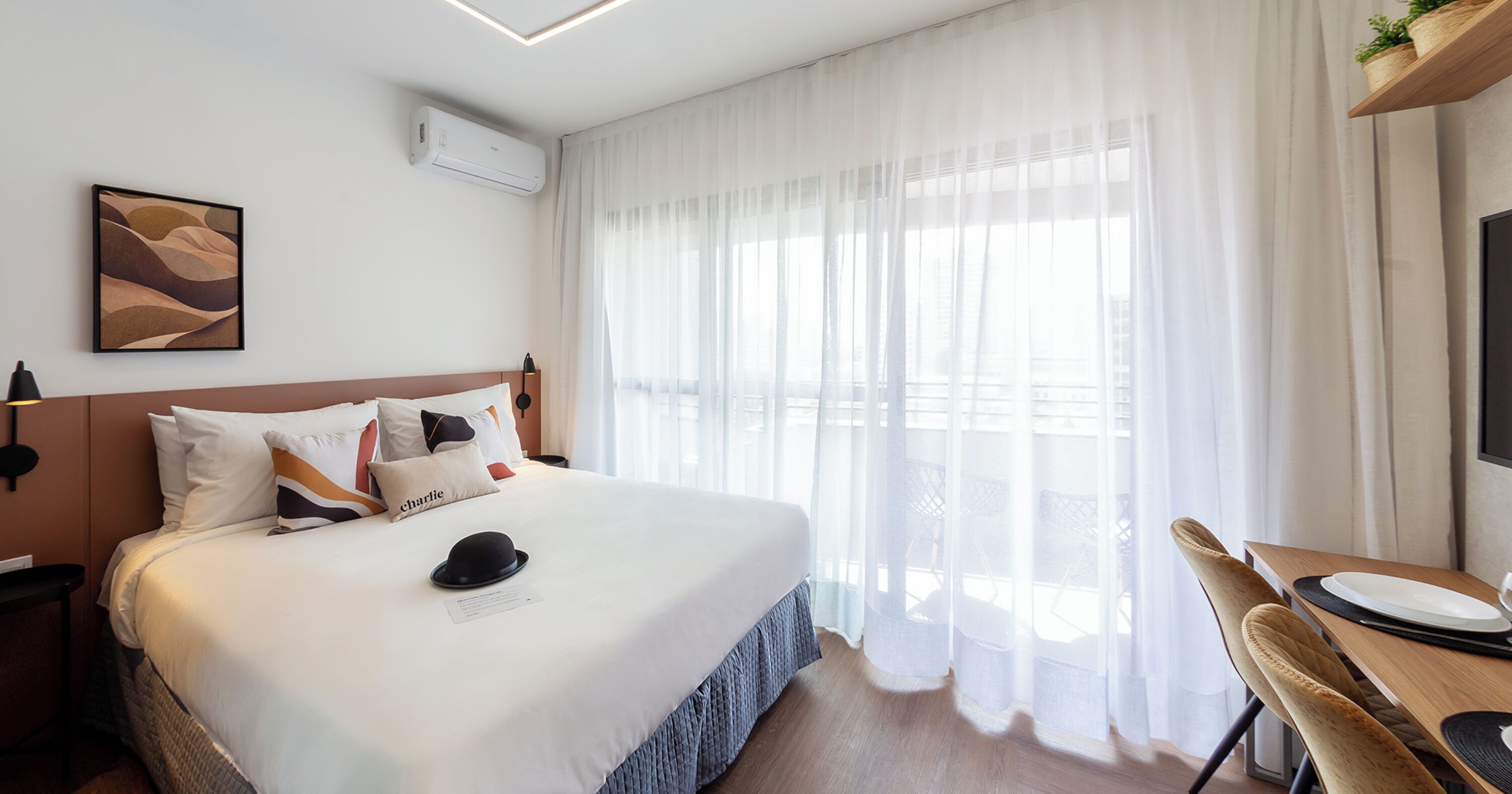 A hotel room with a large bed, a desk with a chair, a window with sheer curtains, and an air conditioning unit on the wall.