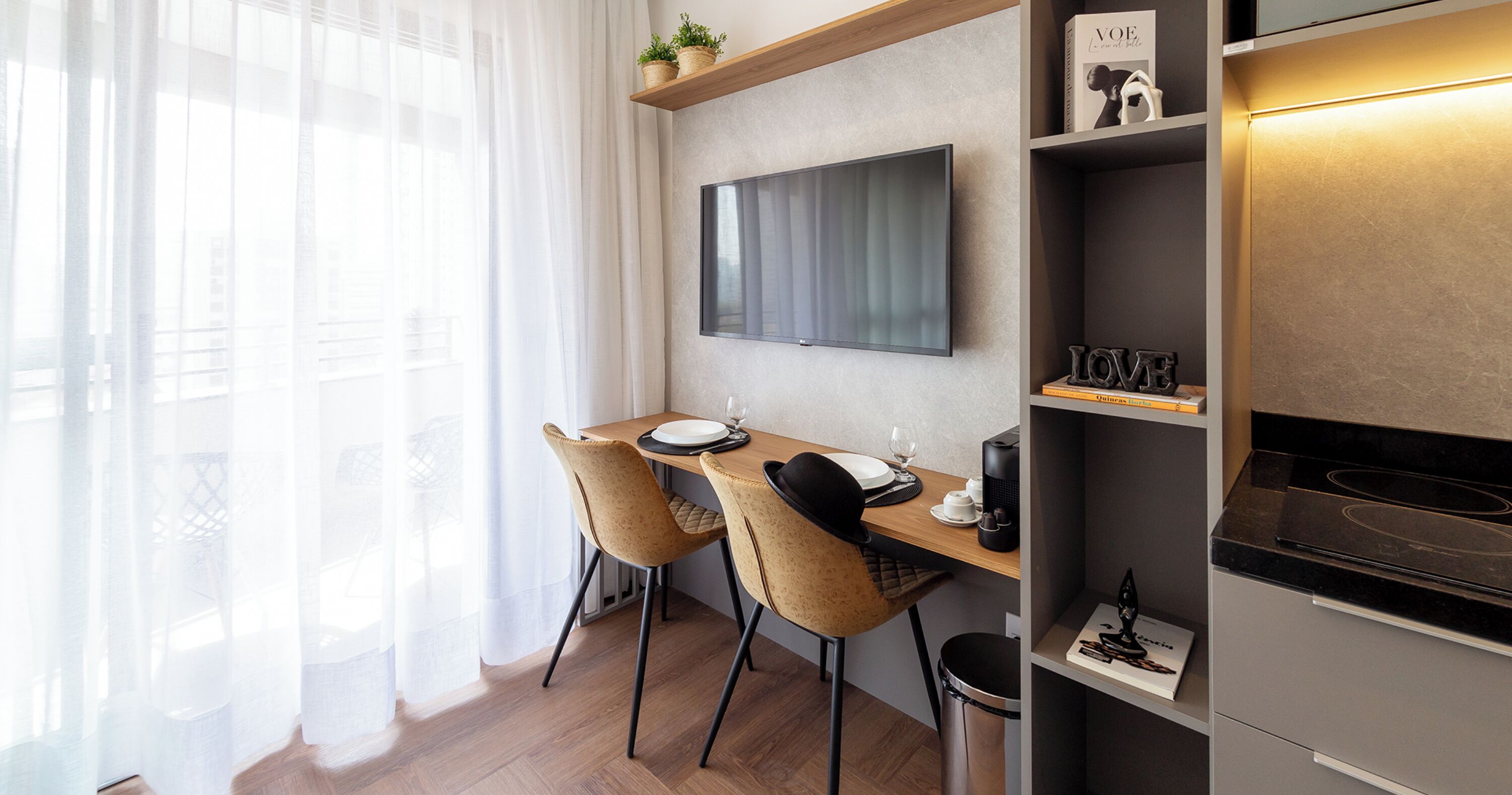 A modern dining area with a wooden table, two chairs, a flat-screen TV mounted on the wall, and a kitchen counter with a stove.
