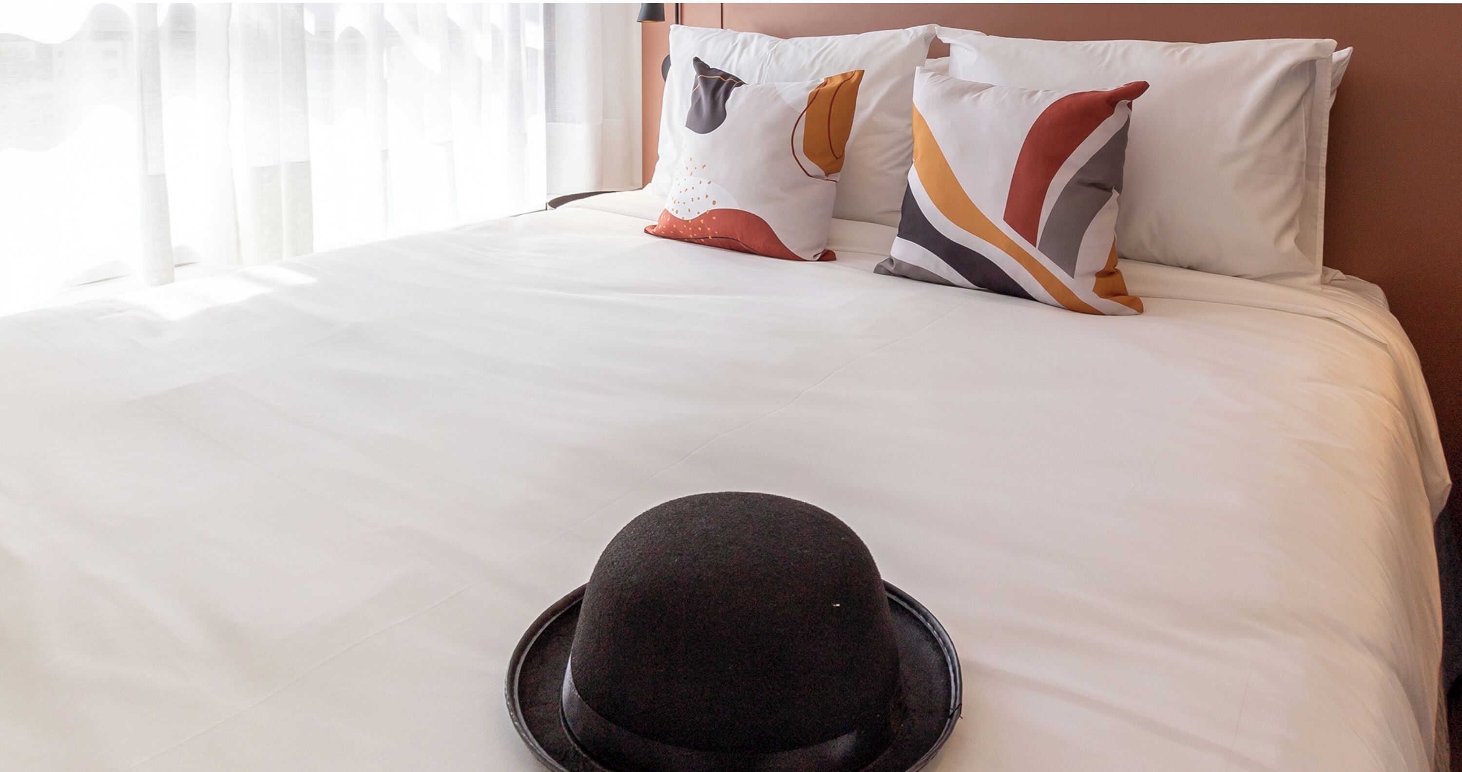 Cama bem arrumada com lençóis brancos, um chapéu preto sobre a cama e duas almofadas com desenhos abstratos.
