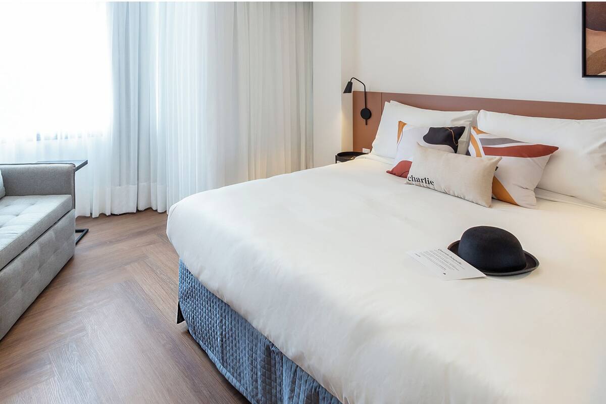 Ein ordentlich bezogenes Bett mit weißer Decke und mehreren Kissen, ein Nachttisch mit Lampe, ein kleiner Topfpflanze und ein Sessel in einem modernen Hotelzimmer.