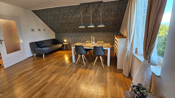 A modern living room with a wooden floor, a sofa, a dining table with chairs, and a kitchen area with a window.