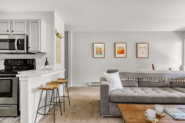 A modern kitchen with white cabinetry, a built-in microwave, and a stove. There is a grey sofa with a white cushion and a wooden coffee table with two white cups.