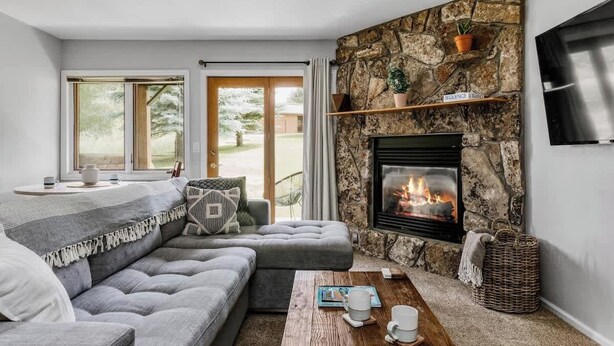 Ein gemütliches Wohnzimmer mit Kamin aus Stein, einem grauen Ecksofa, einem Holztisch und einem großen Fenster mit Blick ins Freie.