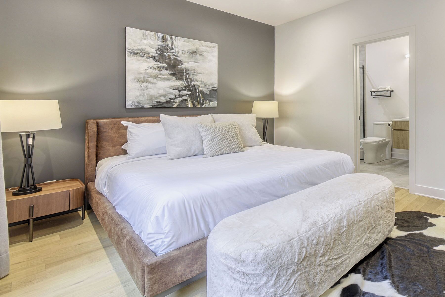 Une chambre avec un grand lit, deux lampes de chevet, une table de nuit, un tableau au mur et une salle de bain visible à travers une porte ouverte.