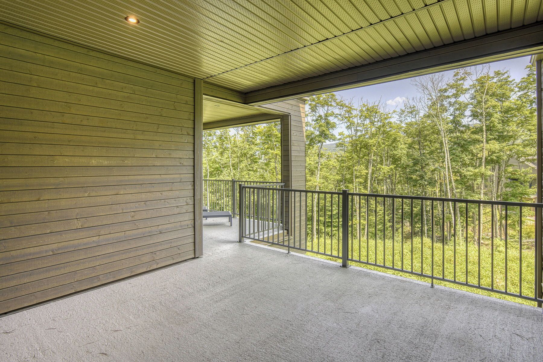 A covered balcony with a concrete floor, metal railing, and a view of a forested area.