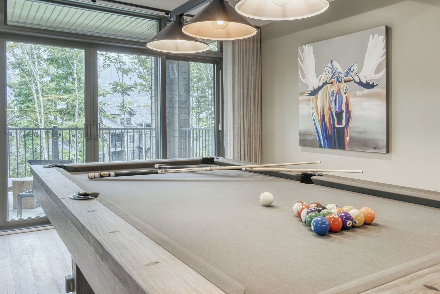 A pool table with pool cues and balls, a painting of a moose, and a view of trees through large windows.