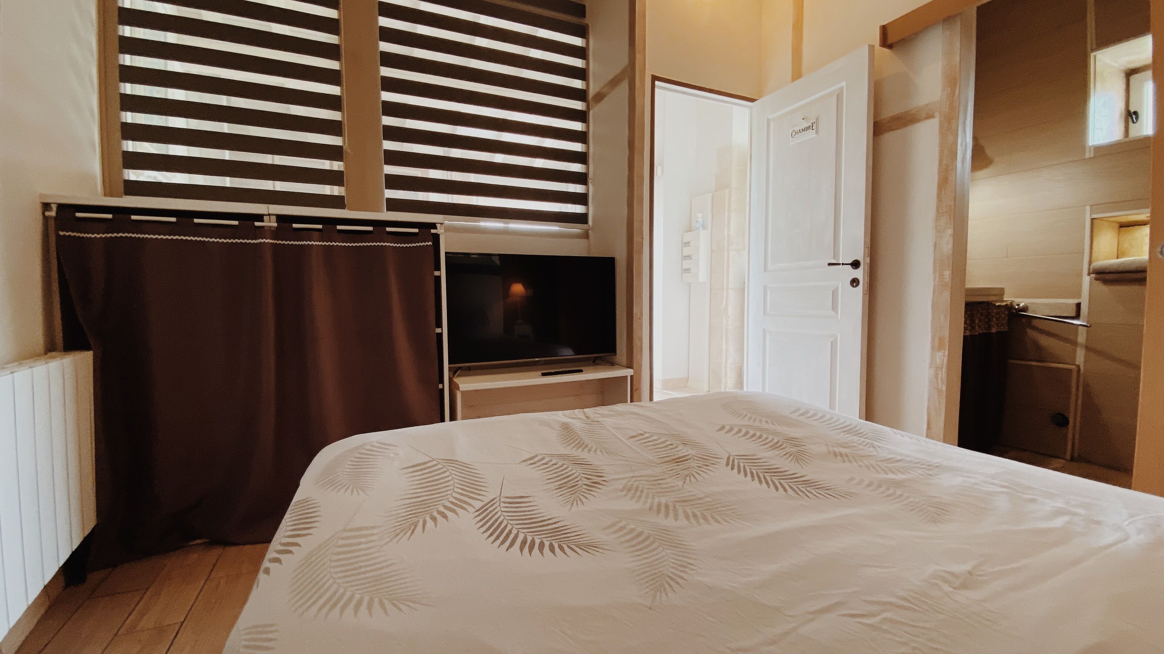 Ein Schlafzimmer mit Bett, Fernseher, Tür und einem Fenster mit Jalousien.