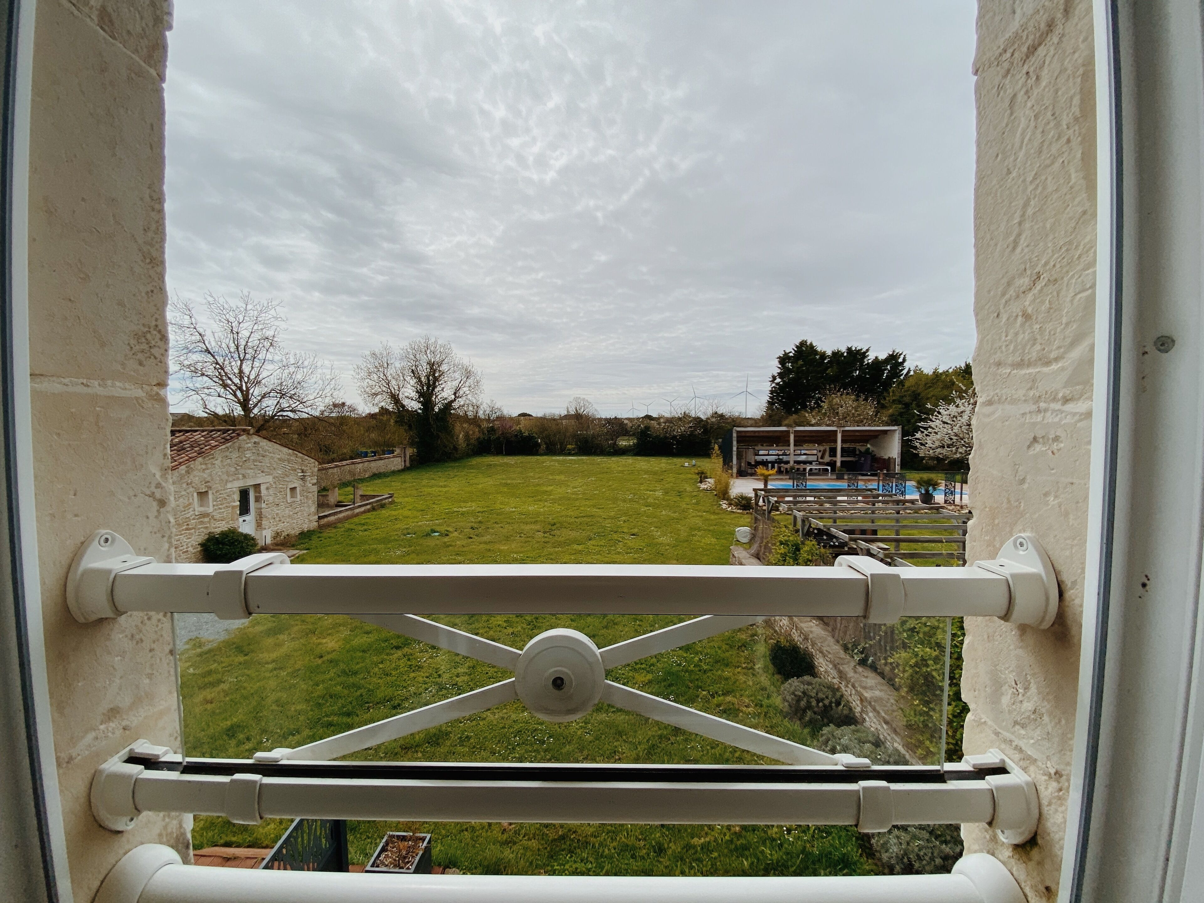 A view from a window with a metal guardrail, a grassy yard, a stone building, and a signboard in the distance.