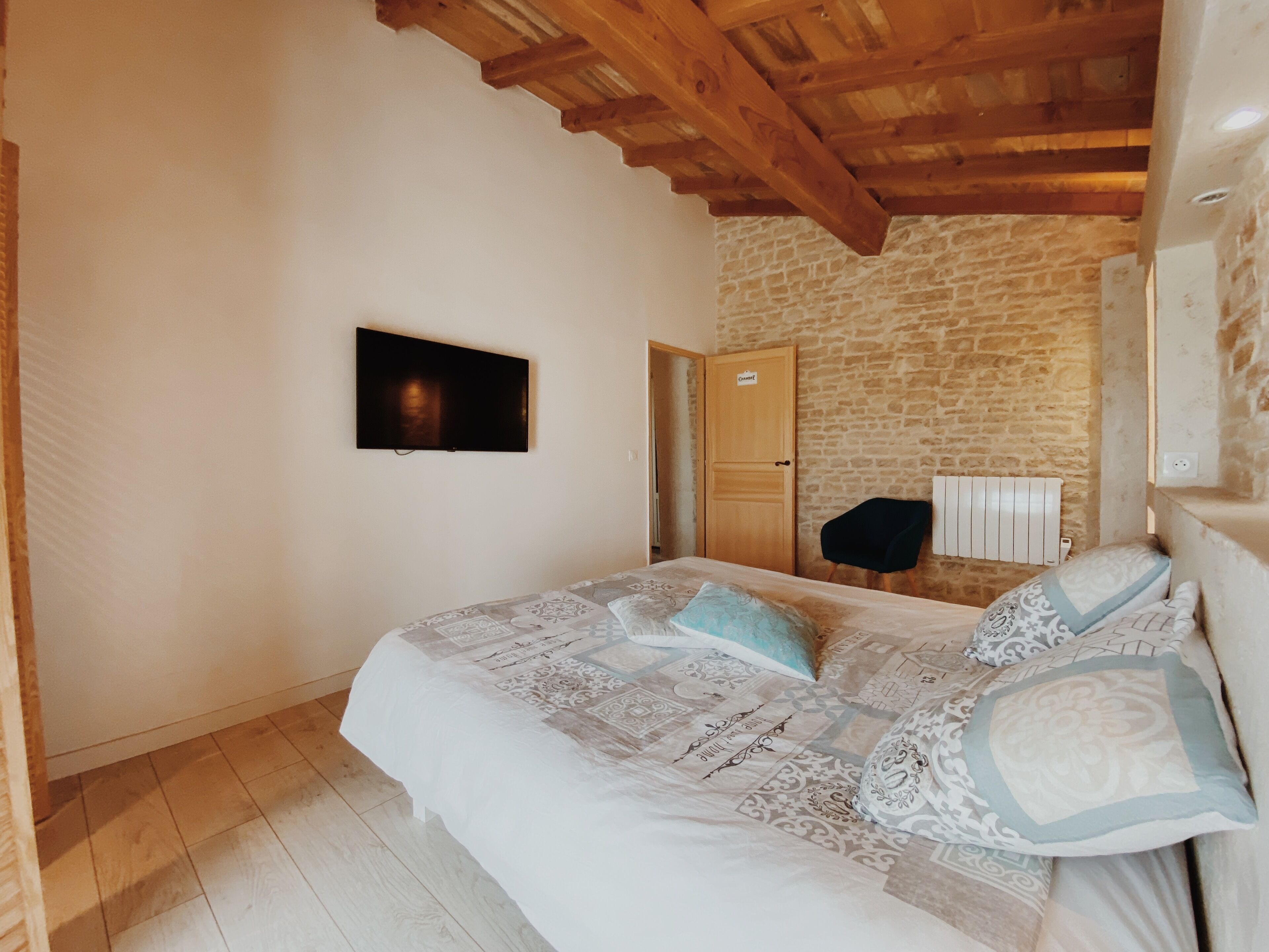 A bedroom with a bed, a television mounted on the wall, a wooden ceiling, and a stone wall.