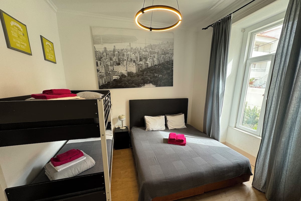 Une chambre avec un lit superposé, un lit simple, un tableau représentant un paysage urbain et une fenêtre avec des rideaux.
