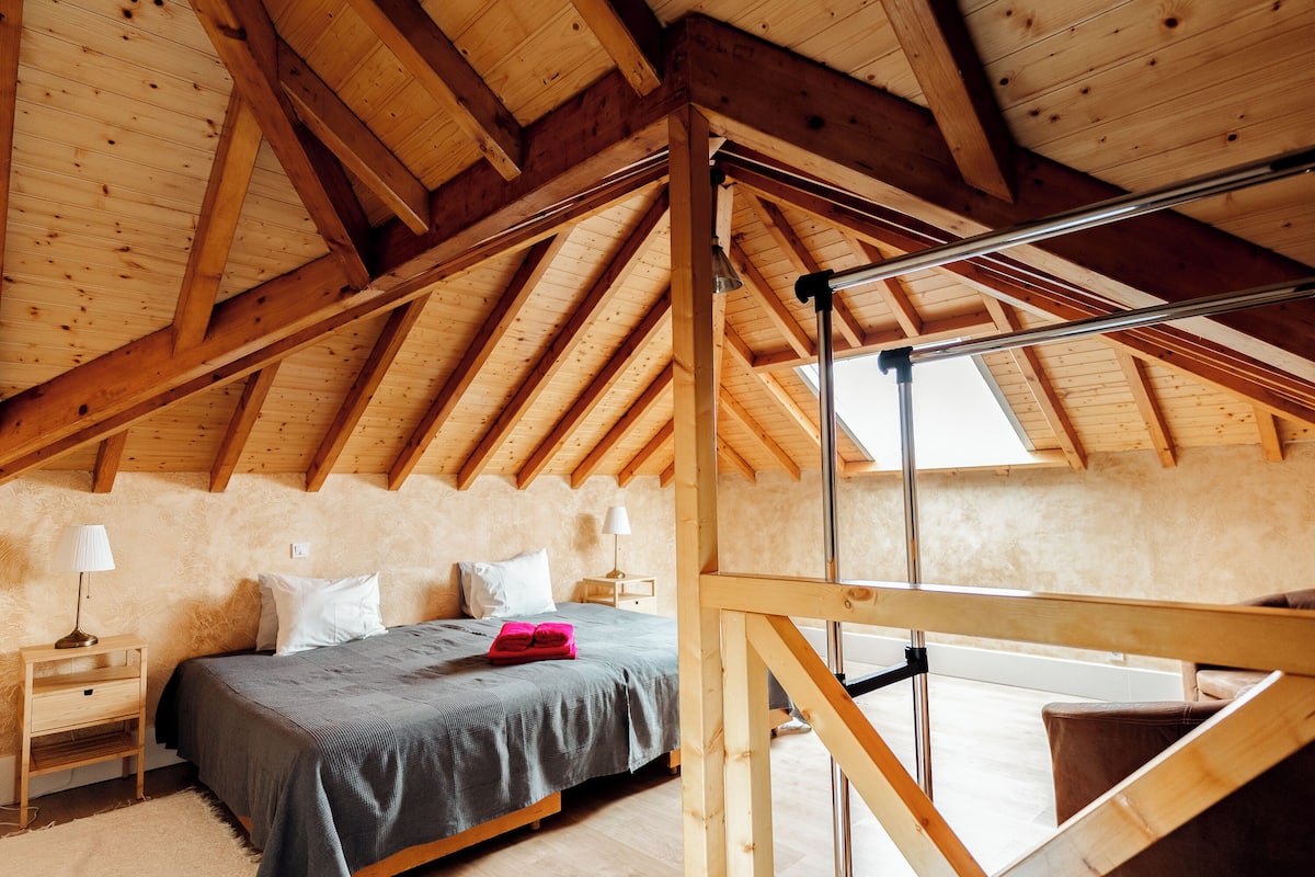 Une chambre avec un plafond en bois incliné, un lit recouvert d’une couverture grise, deux tables de chevet avec des lampes et une fenêtre.