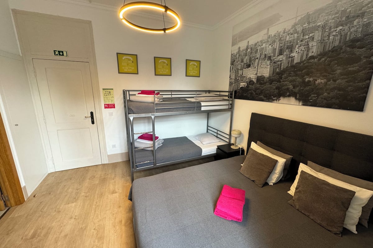 Une chambre d’hôtel avec un lit superposé, un grand lit avec des oreillers, un luminaire fixé au mur et un tableau représentant un paysage urbain.