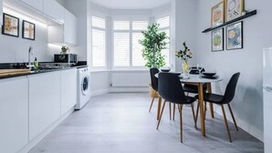 A modern kitchen with white cabinetry, a dining area with a round table and black chairs, and a large window with blinds. - Parkview Place by Patternbricks (Coventry)