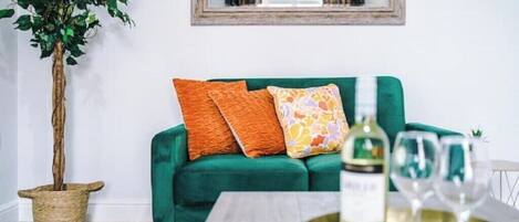 A green sofa with orange and multicolored pillows, a bottle of white wine, and two glasses on a tray.