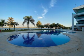 Un hotel moderno con una grande piscina a forma di curva, circondata da palme e lettini.