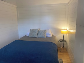 A bed with a blue blanket, pillows, and a bedside lamp in a room with white paneled walls.