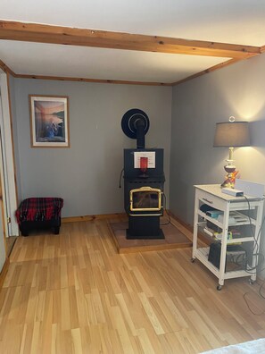 Une pièce avec un plancher en bois, un poêle noir, une étagère blanche où sont posés divers objets, et un tableau encadré accroché au mur.