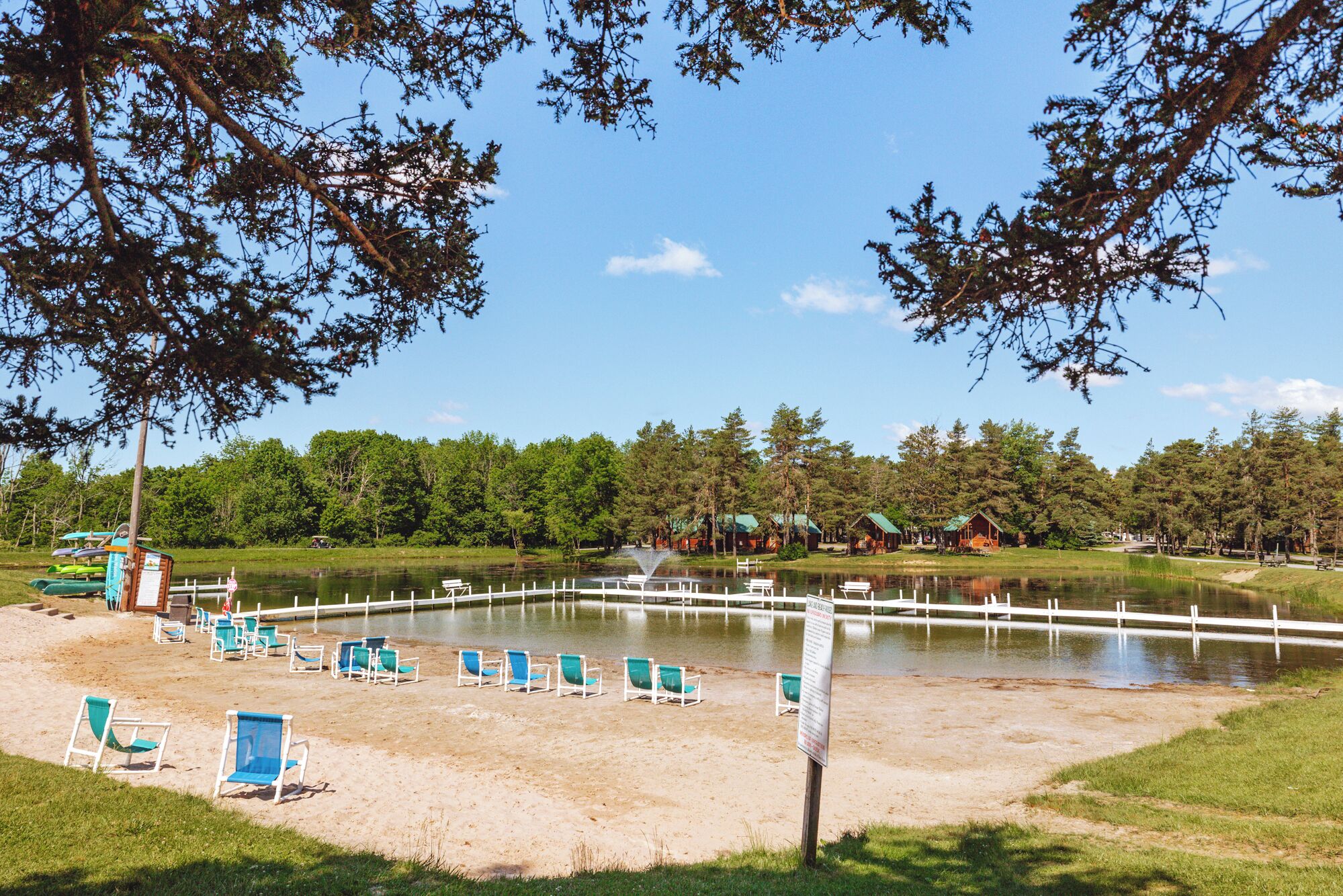 A lakeside resort with a sandy beach, lounge chairs, and cabins in the background.
