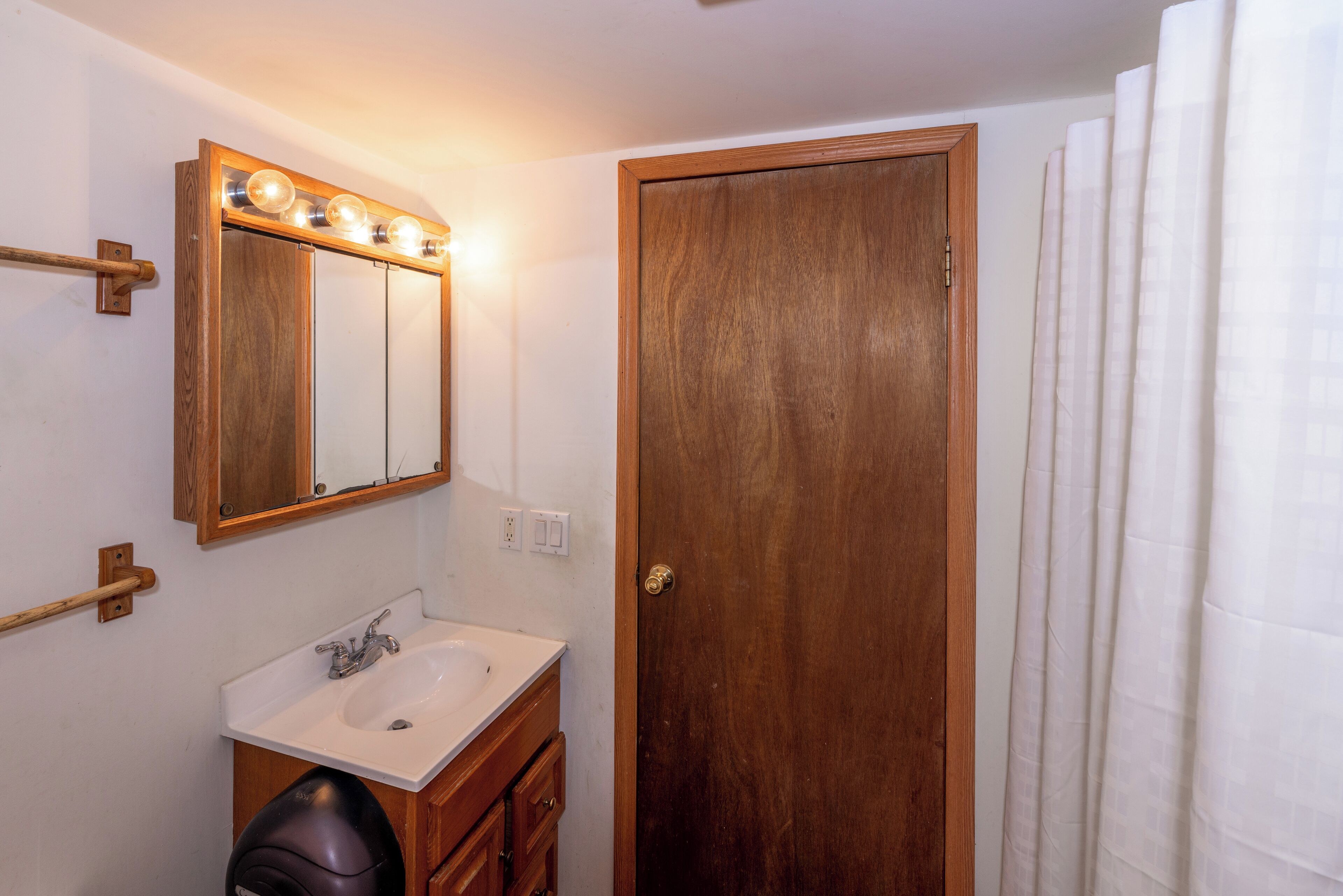A bathroom with a wooden door, a white sink, a mirror with lights, and a towel rack.
