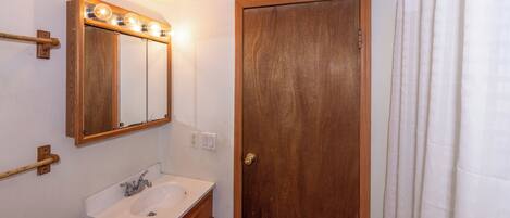 A bathroom with a wooden door, a white sink, a mirror with lights, and a towel rack.
