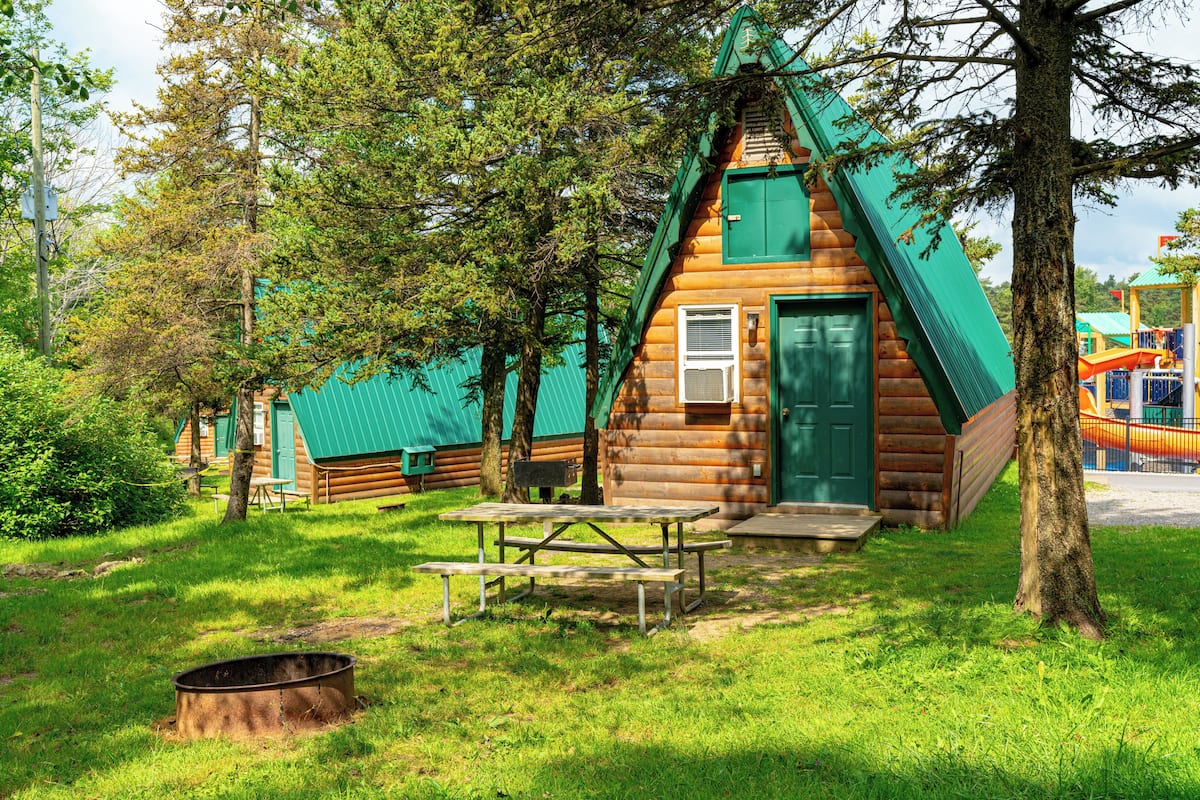 A wooden cabin with a green door and windows, surrounded by trees and a picnic table.