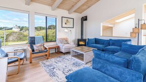 Uma sala de estar aconchegante com um sofá grande, uma mesa de centro e uma lareira. Janelas amplas proporcionam vista para o exterior.