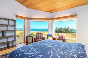 A bedroom with a large bed, a bookshelf, two chairs, and a view of the ocean.