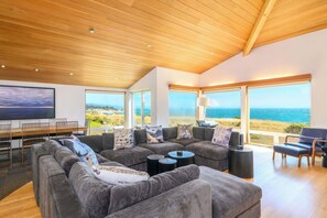 A spacious living room with a large gray sectional sofa, a wooden ceiling, and large windows offering a view of the ocean.
