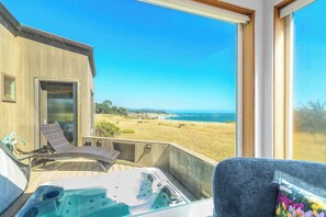 A room with a large window overlooking a beach and ocean, a hot tub, and a chair.