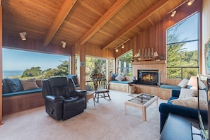 A cozy living room with a fireplace, seating areas, and large windows offering a view of the outdoors. - Viglione | Fireplace, Hot Tub, & Ocean Views (Sea Ranch)