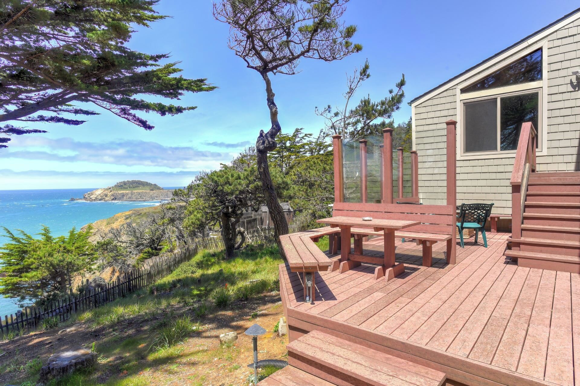 Une terrasse en bois avec une table et des chaises, offrant une vue sur un paysage côtier arboré et sur l’océan.