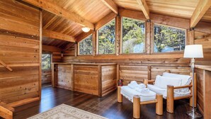 L’intérieur d’une cabane en rondins, avec un lit en bois, une lampe et de grandes fenêtres donnant sur la forêt.