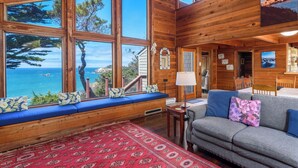 Un chalet confortable doté de grandes fenêtres offrant une vue sur l’océan, d’un coin salon agréable avec un canapé et un banc, et d’un tapis rouge à motifs sur le sol.