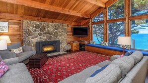 L’intérieur chaleureux d’un chalet avec une cheminée en pierre, un téléviseur et une grande fenêtre donnant sur une zone boisée.