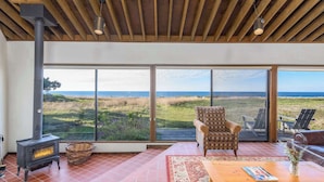 A spacious room with a large window overlooking a grassy landscape and the sea. A traditional wood-burning stove is visible, and there is a wicker basket on the floor. - Sea Pine | Hot Tub with Ocean View (Sea Ranch)