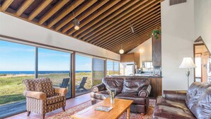 A spacious living area with a large window overlooking a scenic landscape, a wooden ceiling, and a cozy seating arrangement. - Sea Pine | Hot Tub with Ocean View (Sea Ranch)