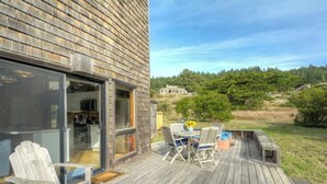 Eine Holzterrasse mit Gartenmöbeln, eine Glastür, die zu einer Küche führt, und Blick auf einen bewaldeten Bereich.