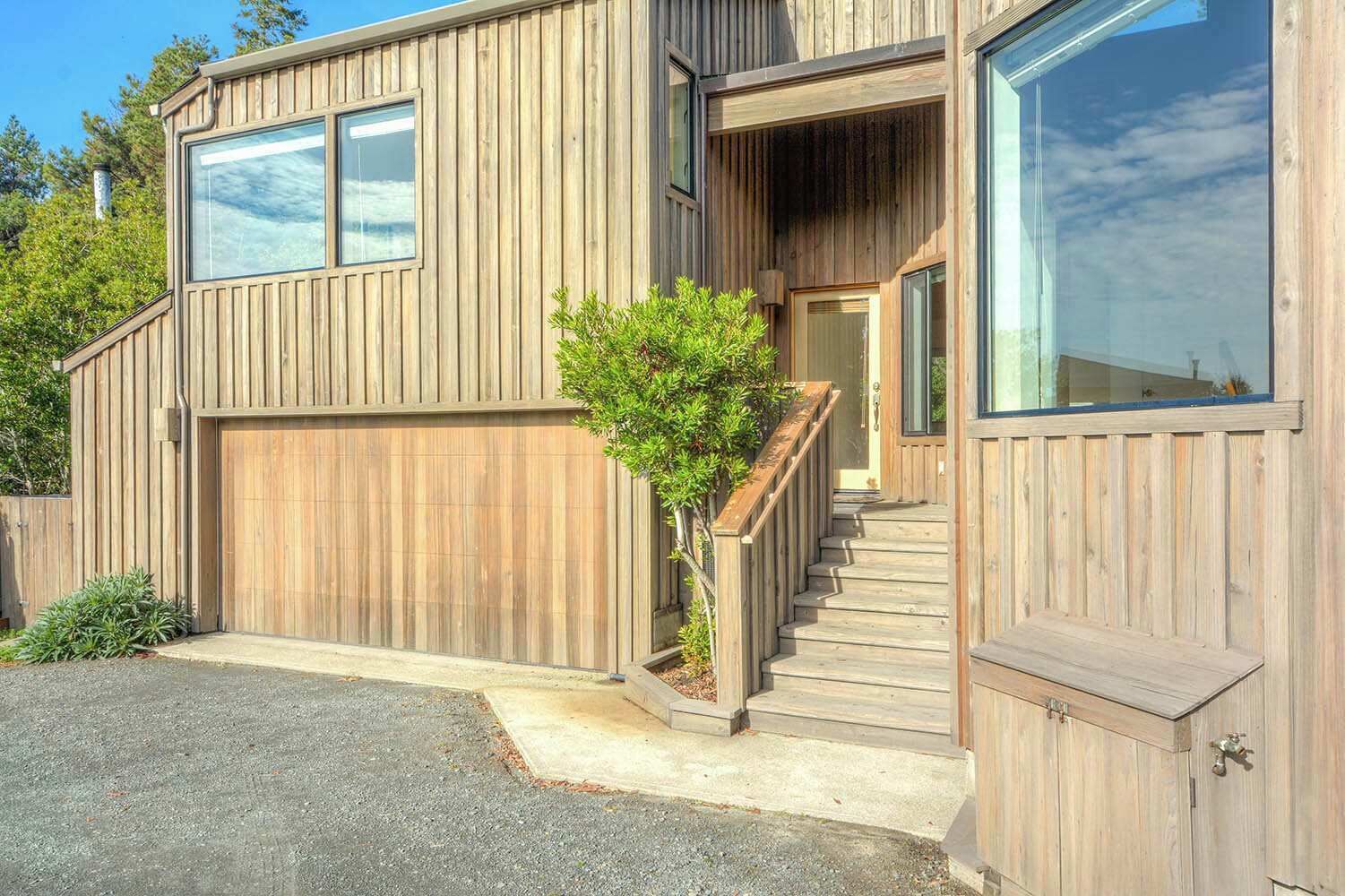A modern wooden house with a garage, a staircase, and a small tree in the front yard.