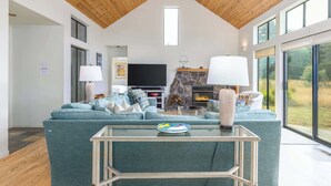 A spacious living room with a high ceiling, a stone fireplace, a large TV, and a view of the outdoors through large windows. - Soak in Ocean Views at Blair w/ Hot Tub! (Sea Ranch)