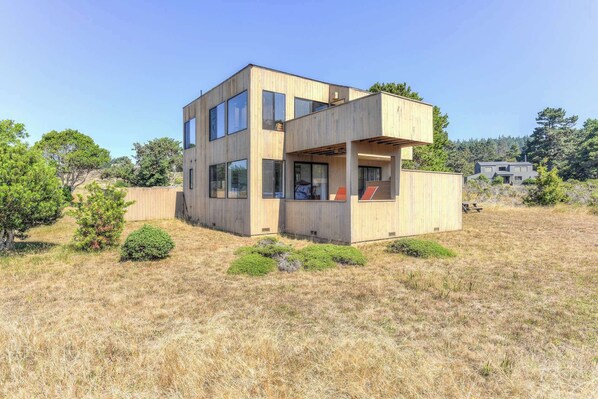 A modern two-story house with large windows and a flat roof, surrounded by dry grass and trees. - Sea Meadow | Panoramic Ocean Views (Sea Ranch)