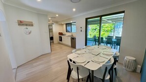 A modern dining area with a table set for six, a kitchen in the background, and a large window with a view of greenery. - Broadwater Beach Cottage (Broadwater)